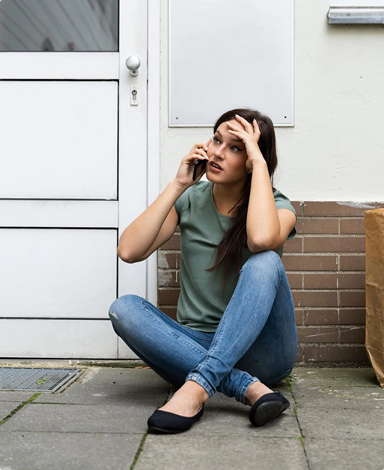 A woman locked out of her house sitting on the ground in front of the front door, calling a mobile locksmith.