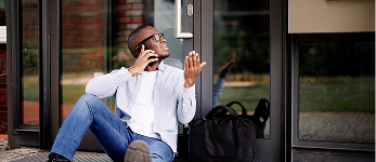 A man sitting on the ground outside of his house, calling a mobile locksmith to unlock the door.